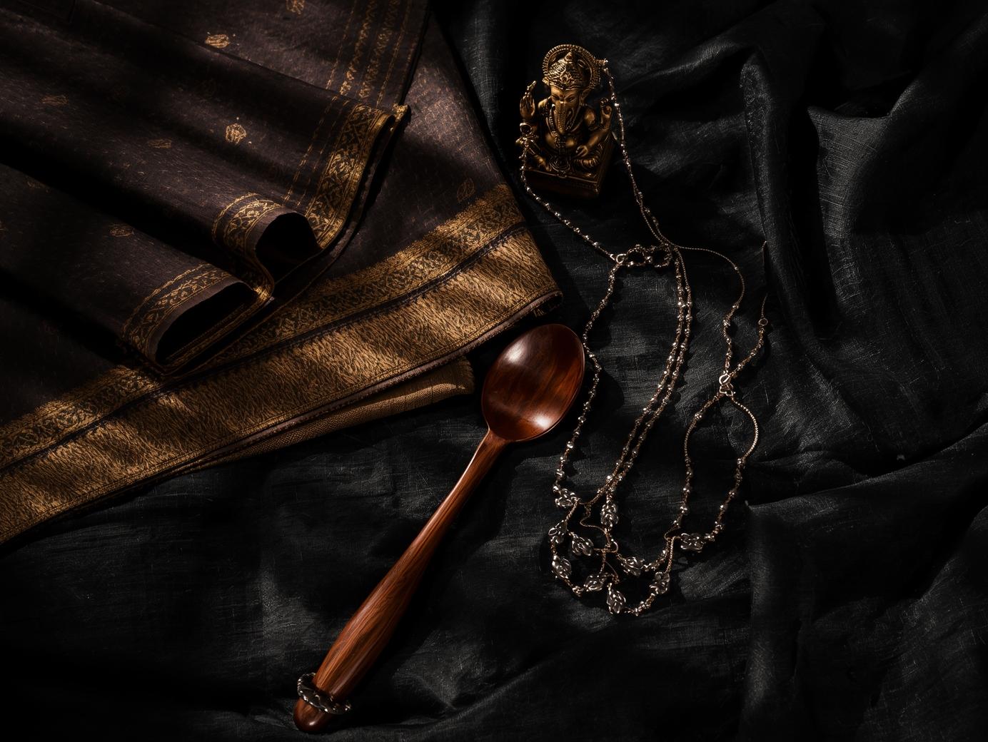 Flat-lay of handcrafted Indian products including a handloom saree, wooden spoon, oxidised jewellery and a bronze idol displayed on black fabric – ManiCrafted collections.